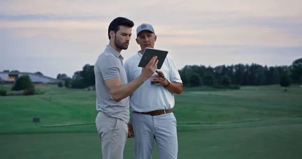 Two golfers looking tablet watching tournament game video at golf course sunset. Team players surfing internet information on digital device tab. Wealthy men read news on pad. Hobby technology concept