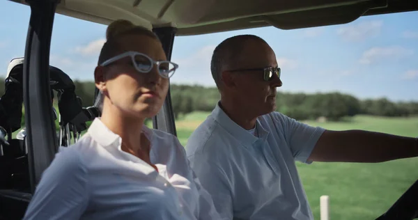 Two players sit golf cart on active summer weekend. Couple enjoy country club leisure close up. Professional team golfers spend time together on course landscape view. Golfing luxury hobby concept.