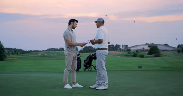 Two businessmen stand outside on golf course. Sportsmen sign paper on field country club. Wealthy rich golfers team talking discussing agreement together at sunset nature. Luxury hobby partner concept