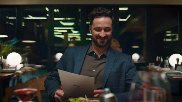 Smiling man looking menu on restaurant date. Happy guy sitting at fancy dinner table. Gentleman choosing tasty food in luxury cafe. Pov date with handsome businessman in eating place. Dining concept.