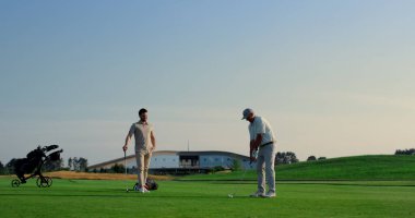 Golf takımı yeşil çim sahası kulübünde spor yapıyor. İki golfçü gün batımında topa vuruyor. Oyuncular birlikte dışarıda lüks bir hafta sonu geçirmenin keyfini çıkarıyorlar. Erkekler çim biçme yerinde eğleniyor. Etkin konsept.