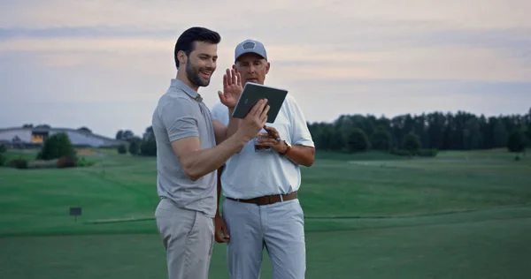 Wealthy men video call tablet device on golf course. Two rich friends talk online outside. Smiling golfer partners using digital tab saying hi on conference meeting. Modern golfing technology concept.