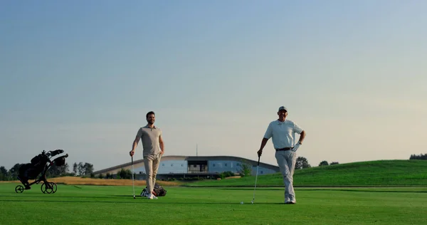 Two men enjoy golf on fairway field club. Golfing team practicing play sport on sunset course. Focused coach group hitting ball using putter clubs outside. Golfers enjoy game on nature. Active concept
