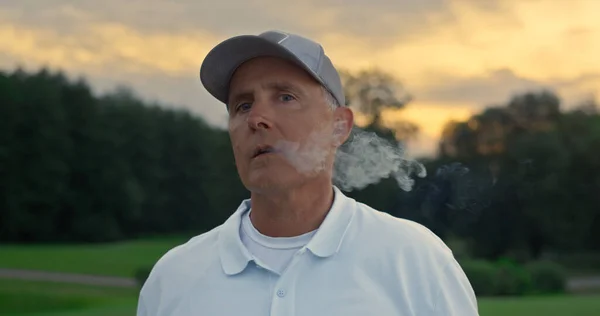 Senior golf player smoking cigar looking camera on sunset field course fairway. Close up wealthy relaxed golfer enjoy summer evening at country club. Old businessman wear sportswear. Luxury concept.