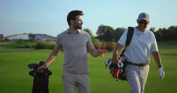 Successful golfers smiling discuss golf on course. Two men enjoy sport action on fairway. Joyful golfing players walk in countryside club on active summer weekend vacation. Friendship activity concept