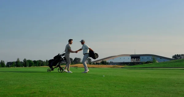 Golf players meeting together on course field. Golfing team talking discussing sport game outside. Two men golfers shaking hands in countryside club. Friends enjoy weekend activity. Leisure concept.