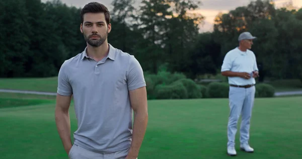 Focused model enjoy golf on sport team outdoors. Two players standing on green course. Attractive bearded guy posing at sunset fairway. Group men rest at luxury country club. Relax golfing concept.