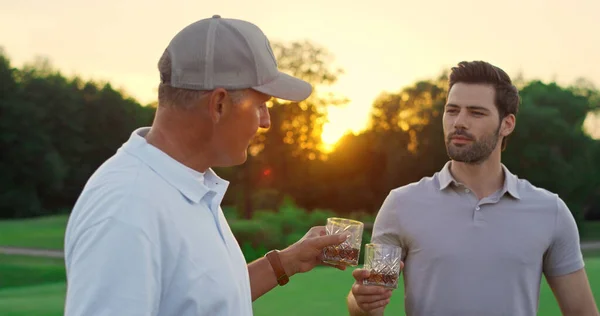 Two golfers chatting together at green course. Golf team enjoy drink whiskey outside at golden sunset. Professional players talking discuss golfing game at country club. Luxury resort summer concept.