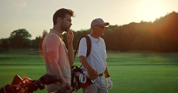 Rich men playing golf on sunset summer course. Two players enjoy sport on field. Professional golfers team looking distance in countryside club. Active group stand in sportswear. Recreation concept.