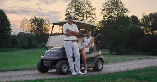 Couple posing golf cart outside. Two golfers take clubs sport equipment on sunset field. Wealthy married lovers enjoy relax activity on weekend. Golfing team in driver seat. Active leisure concept.