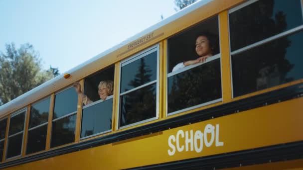 两名多族裔学童从黄色校车窗口往外看 快乐的年长者男孩挥动着父母的手 梦想中的女孩站在校车里笑着 快乐的青少年准备学习 — 图库视频影像