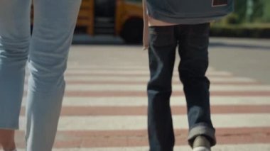 Anne oğul bacakları yaya geçidinde yürüyor. Aile el ele tutuşuyor. Bilinmeyen anne, küçük çocuğa okula kadar eşlik ediyor. Çocuk okul çocuğu sırt çantasını sarı okul otobüsüne taşıyor güneşli yaz sabahında..