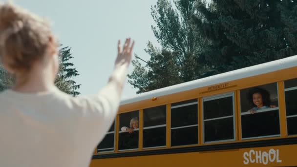 可爱的妈妈乘黄色校车向小儿子挥手 孩子们在阳光灿烂的早上上学 金发女子给小学生送去了一个飞吻 有爱心的妈妈站在街上等公共汽车离开 — 图库视频影像
