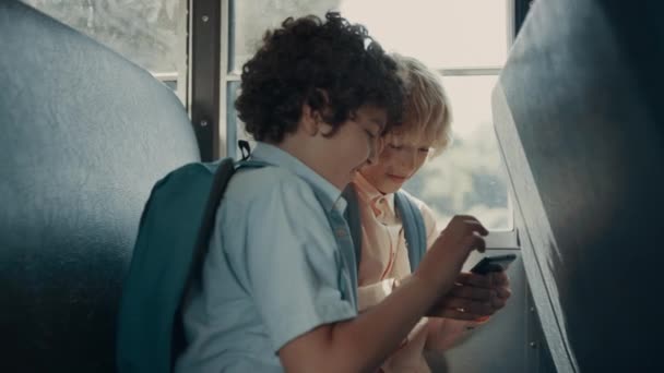 電話に座ってスクールバスでゲームをプレイする2人の興奮した小学生 毎日の旅行で現代のスマートフォンを使用して陽気な男の子 幸せな乗客は家に帰る途中でサーフィンのウェブを楽しむ — ストック動画