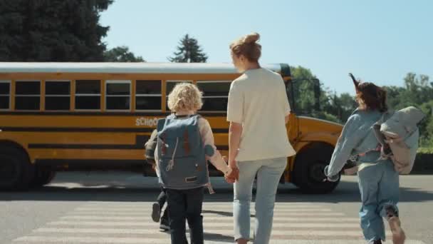 妈妈牵着男同学走向黄色的校车 精力充沛的十几岁的学生走过马路 年轻的妈妈陪着卷曲的儿子去阳光灿烂的早上上学 青少年人群跑上了学术穿梭车 — 图库视频影像