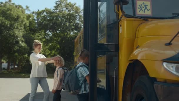 妈妈说再见了 可爱的小儿子站在黄色的公共汽车旁 不同的学生登上校车 快乐的妈妈把孩子卷曲的头发弄乱了 笑着男孩走进了学校的穿梭车走开了 学习概念 — 图库视频影像