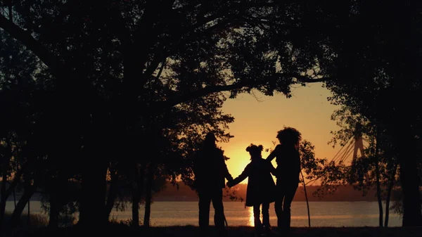 Happy family silhouette walking sunset park together. Beautiful golden sunlight. Unrecognized parents holding hands enjoy leisure time with daughter at river shore. Unconditional love devotion concept