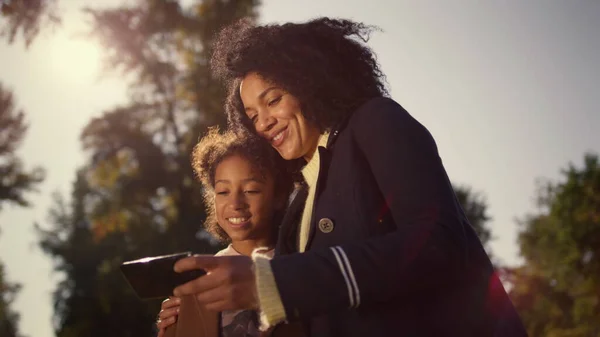 Happy mother daughter selfie posing in golden sunlight. Joyful time together. Attractive african american woman holding smartphone make picture with smiling kid. Cute family enjoying moment sunny day