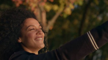Kadın elini güneş portresine doğru uzatıyor. Aile şehir yoğunluğundan kaçtı. Çekici Afro-Amerikan kıvırcık kadın sonbahar parkında sıcak altın güneş ışığının tadını çıkarıyor. Gülümseyen kız dinleniyor..