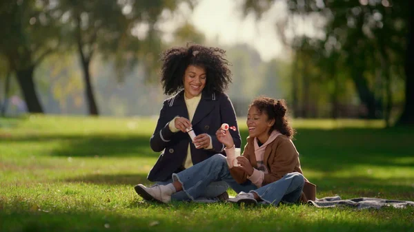 Happy daughter blowing soap bubble sitting in sunny park. Joyful family weekend. Smiling african american girl having fun playing with pretty curly mother on picnic. Loving parent laugh enjoy kid time