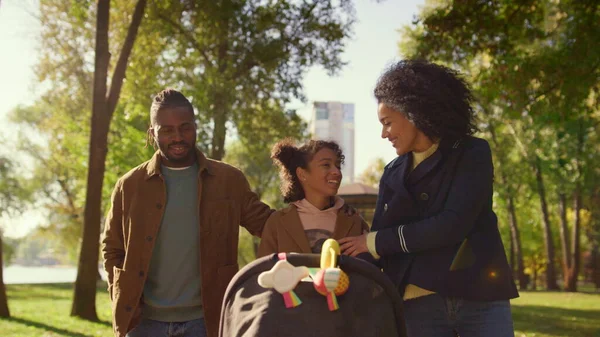 Happy family talking walking together in sunny park. Joyful family weekend. Smiling african american parents loving mother father enjoying time with kids. Caring sister pushing baby stroller walkway.