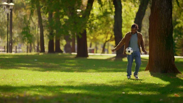 Happy father playing football in sunny park alone. Smiling player having fun. Focused joyful african american man enjoy warm weekend practicing soccer ball tricks. Active hobby adult lifestyle concept