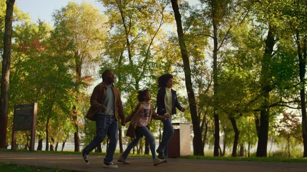African american family walk autumn park together. Happy kid hold parents hands. Joyful girl making jump enjoying evening leisure time with mother father in city garden. Carefree childhood moments.