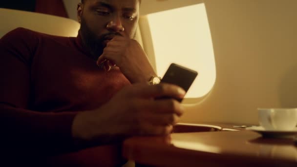 スマートフォンのメールをチェックするビジネスマンに焦点を当て 携帯電話を持っている手を閉じる アフリカ系アメリカ人の男性Ceoが飛行機でプロジェクトデータソーシャルメディアのニュースを読んでいる 出張のコンセプト — ストック動画