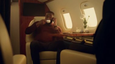 İş seyahatinde konuşan Afro-Amerikan akıllı telefon. Keyifli bir adam birinci sınıf bir uçakta dinleniyor. Neşeli, başarılı, siyah saçlı iş adamı kahve içiyor ve partneriyle iletişim kuruyor..