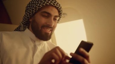 Uçak penceresinde başarılı sörf yapan adam. Yakın plan. Arap akıllı telefonu özel jette sosyal medya içeriğinin keyfini çıkarıyor. Neşeli sakallı adam haberleri okuyor e-postaları kontrol ediyor. İş seyahati kavramı