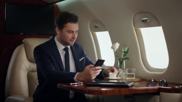 欧洲商人在飞机旅行中输入电话 自信的经理在豪华喷气式飞机上喝咖啡 成功人士穿着西服发短信上网 快乐绅士检查电子邮件使用智能手机 — 图库视频影像