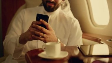 Gülümseyen adam uçakta mesaj atıyor. Jette akıllı telefonu tutan yakın el. Müslüman kıyafetiyle cep telefonu yazan neşeli Arap işadamı. İş gezisine odaklanmış sakallı yolcu.