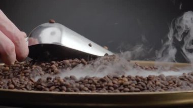 Kavrulmuş kahve çekirdekleri kepçeyle kepçeyle kaplanıyor. Kimliği belirsiz bir adam elinde kepçeyle kızartma makinesinde aromatik tahıl karıştırıyor. Kahverengi kahve çekirdeklerinin üzerine ağır çekimde hafif buhar yükseliyor..