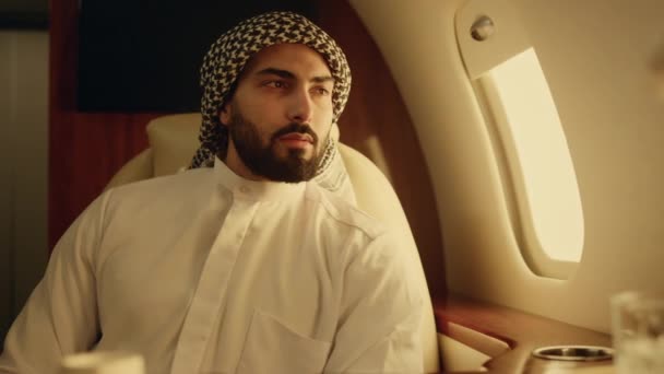 イスラム教徒の服の若いアラビア語を探してカメラを閉じます 笑顔の男は企業旅行のジェットウィンドウで休む 豪華な民間航空機を旅する裕福な思考ビジネスマン ビジネスライフスタイルコンセプト — ストック動画