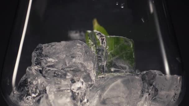 ミントのアイスカクテルを閉じます 緑色の葉は冷たいガラスの中に落ち 氷塊は黒い背景の超遅い動きをします 新鮮な食材のコンセプトでパーティー飲料を準備する — ストック動画