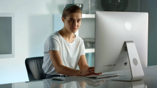 Smiling woman using desktop computer at home office. Corporate worker lifestyle. Serious successful businesswoman typing looking in camera. Confident freelancer creating article at remote workplace.