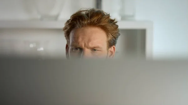 Focused businessman looking computer monitor at home remote workplace closeup. Ginger business analyst professional project manager check project data. Serious freelancer surf behind defocused monitor