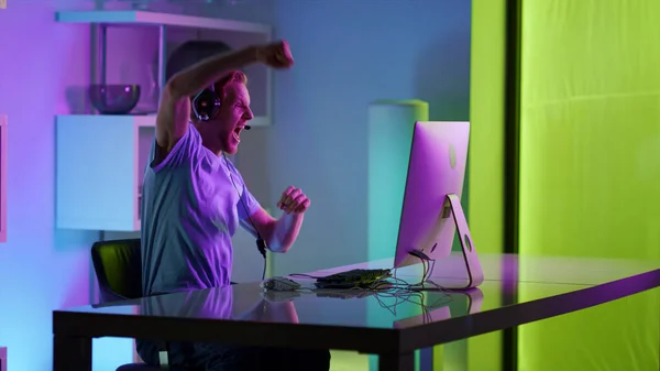 Excited man celebrating gaming tournament victory in neon cyberspace at home. Overjoyed ginger guy shout making yes gesture enjoy cyber triumph. Happy gamer winning computer game resting on weekend.
