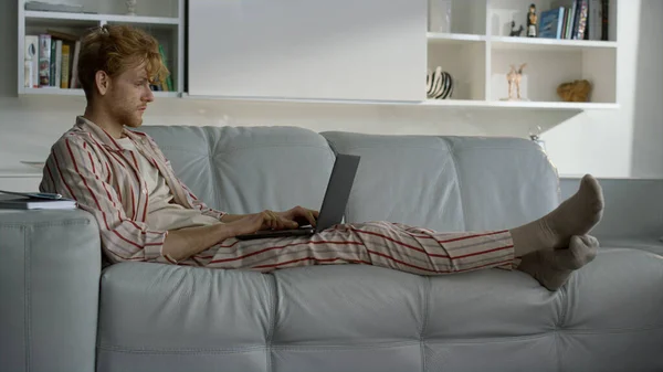 穿着睡衣在家里办公室工作的人 重点姜汁学生使用笔记本电脑在沙发上休息 认真的自由职业者在遥远的工作场所上网浏览手表 检疫生活方式概念 — 图库照片