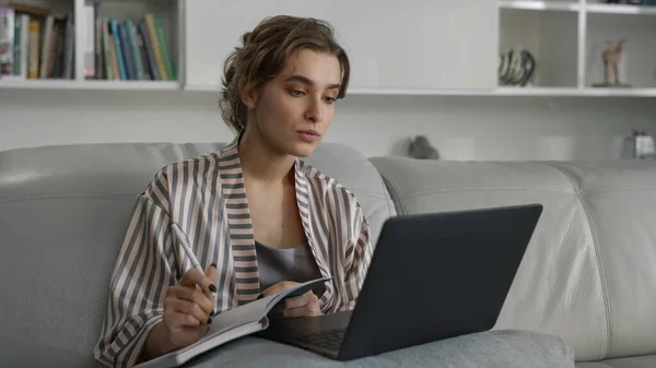 Smiling girl studying home watching online university class in pajamas closeup. Joyful beautiful manager making notes attending training on weekend. Woman use laptop analyzing data at remote office