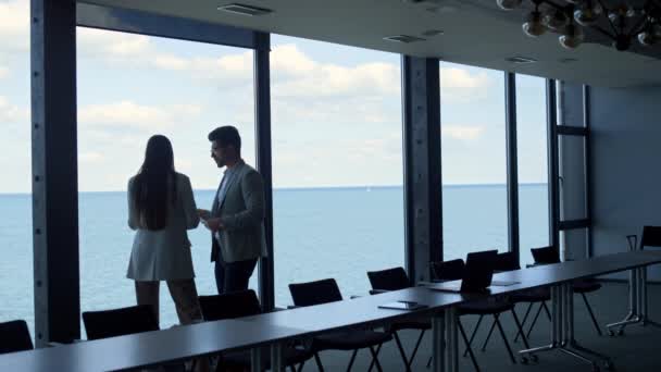 パノラマウィンドウでビジネス取引を議論する2人の同僚 財務責任者は海の眺めでプロジェクトの書類をチェックする ミレニアルCeoの投資家が会議室でビジネスウーマンと会話 — ストック動画