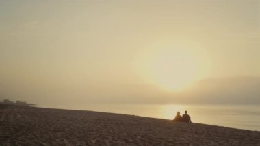 Sahilde gün batımını izleyen sevimli bir çift. Geniş açılı erkek arkadaş ve kız arkadaşın denizde buluşması. Arka plan dostları okyanus kıyısında birlikte dinleniyorlar. Gün doğumunda deniz manzarası. Boş zaman konsepti