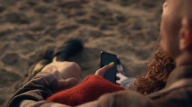 Tanınmayan bir çift kumsalda tatilin tadını çıkarıyor. Yakın plan genç bayan elinde akıllı telefon kullanıyor. Sevgi dolu erkek arkadaş kucaklayan kız arkadaş. Mutlu aşıklar gün batımını birlikte geçiriyorlar. Aşk konsepti
