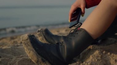 Tatilde sahilde oturan şık bir kız. Cep telefonu tutan tanınmayan bir kadın. Sıradan botlar giyen hippi kadın bacakları. Kumda deri ayakkabılar. Okyanus dalgaları kıyıya vuruyor..