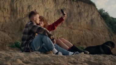 Sevecen bir çift, Sunset Beach 'te akıllı telefondan selfie çekiyor. Mutlu kadın ve adam cep telefonunu kontrol ediyor. Köpekli genç aşıklar kumlu sahilde oturuyorlar. Neşeli hipsterler kucaklaşıyorlar