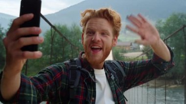 Dağlarda cep telefonu kamerasıyla heyecanlı bir yürüyüşçü görüntüsü. Duygusal hipster el sallamasını cep telefonuyla selamlayın. Doğa manzarasında blog yazan mutlu kızıl adam. Seyahat tatili kavramı