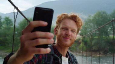 Talking tourist using smartphone make video call on summer mountains. Close up smiling traveler gesture hand at mobile phone camera online. Redhead hipster make inspiring blog on bridge. Trip concept.