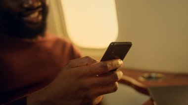 Uçak yolculuğunda internette sohbet eden olumlu bir adam. Akıllı telefonu yakından tutan eller. Mutlu Afro-Amerikan yolcular iş seyahatlerinde uygulama iletişimiyle eğleniyor. Mesajları okuyan mutlu bir adam..