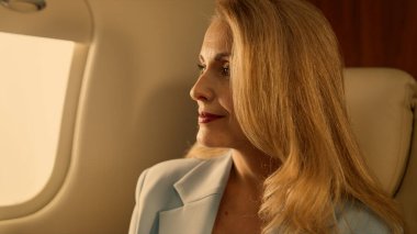 Passenger looking airplane window in golden light closeup. Corporate trip on jet. Attractive confident blonde businesswoman relax in luxury first class. Elegant rich woman thinking traveling in suit clipart