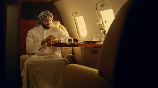 Muslim businessman enjoying airplane trip. Successful man checking hand watch looking jet window. Smiling handsome guy travel luxury private jet in traditional clothing. Wealthy company owner concept